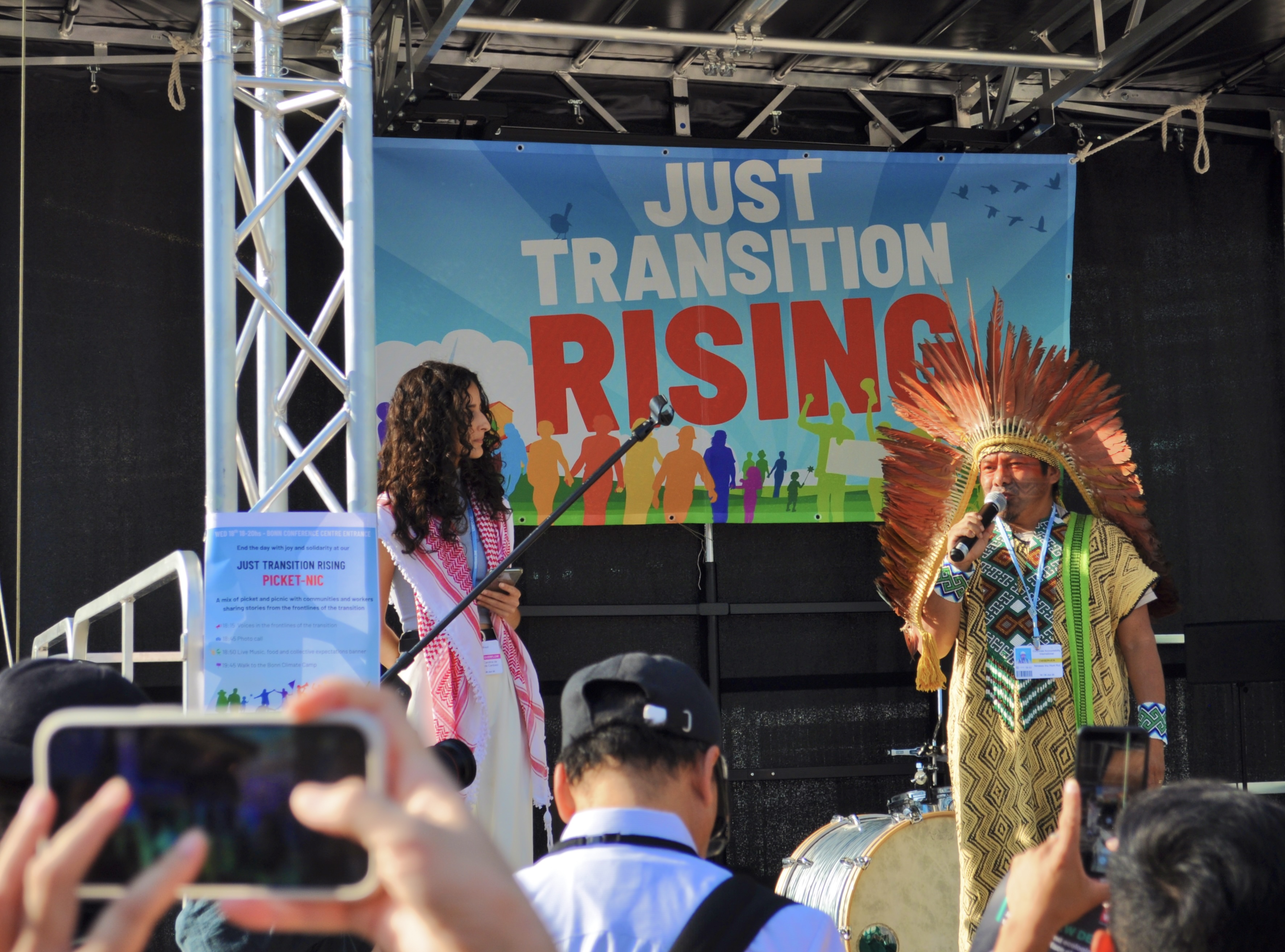 [13] Chief Ninawa Inu Huni Kui, leader of the indigenous Huni Kuin people of Brazil, speaks at the Just Transition Rising protest. A fellow activist stands beside the Chief to translate his speech into English. Chief Ninawa said, "In my country, our land is under constant threat: up for auction for oil and gas, and hydroelectric dams and mining. Many times, it is done in the name of the green economy. [But] what they call innovation, we call repeating old costumes and ways. For my people, the true just tran